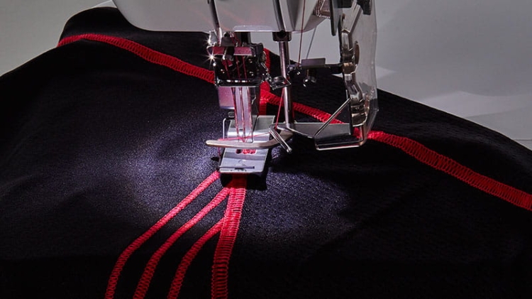 Renderka Brother CV3550 w zbliżeniu, pokazująca igłę i stopkę pracującą na ciemnym materiale z czerwonymi przeszyciami. Widoczna jest też lampa robocza oświetlająca obszar szycia.