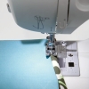 Maszyna do szycia Singer 3342 Fashion Mate, uchwycona z bliska, pokazuje igłę i stopkę szyjącą, pracującą nad niebieskim materiałem z zielonym wzorem. Widoczny panel sterujący z przyciskami i oznaczeniami.