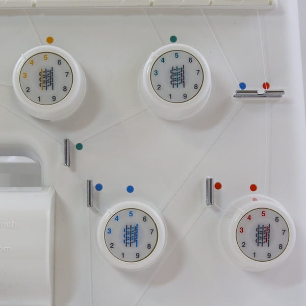 Panel kontrolny owerloka JUKI MO-644D z czterema białymi pokrętłami do regulacji naprężenia nici, z wyraźnymi numerami i kolorowymi oznaczeniami. Widoczny uchwyt oraz elementy do mocowania nici na białym tle.