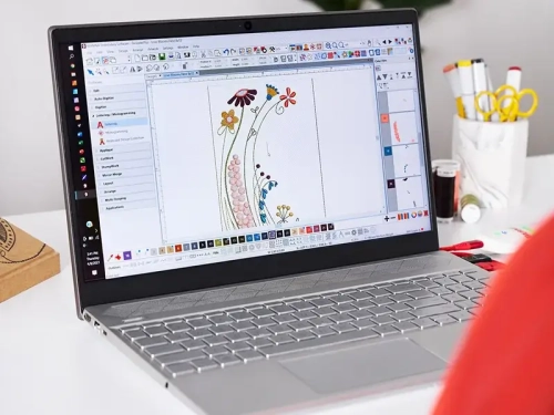Oprogramowanie Bernina V9 Creator na laptopie, wyświetlające projekt haftu z kolorowymi kwiatami na białym tle. Widziane z przodu, na biurku z akcesoriami do szycia w tle, w tym nożyczkami i szpulkami nici.