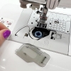 Maszyna do szycia Brother NV15, widok z bliska na mechanizm nawlekania nici. Widać bębenek z niebieską nicią i elementy instrukcji na obudowie. Ręka z fioletowym manicurem trzyma nić, a obok leży przezroczysta osłona z otworami.