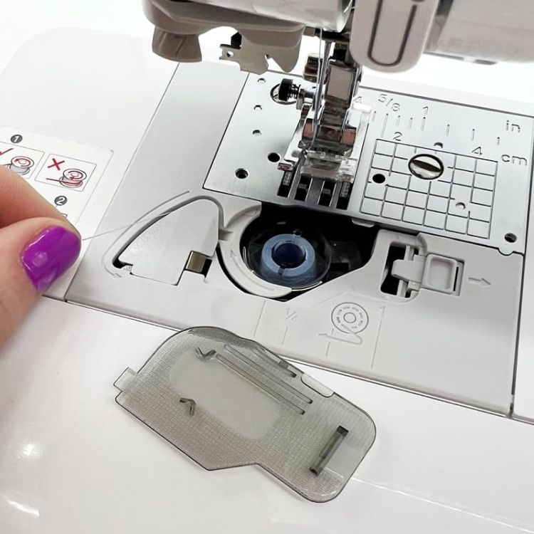 Maszyna do szycia Brother NV15, widok z bliska na mechanizm nawlekania nici. Widać bębenek z niebieską nicią i elementy instrukcji na obudowie. Ręka z fioletowym manicurem trzyma nić, a obok leży przezroczysta osłona z otworami.