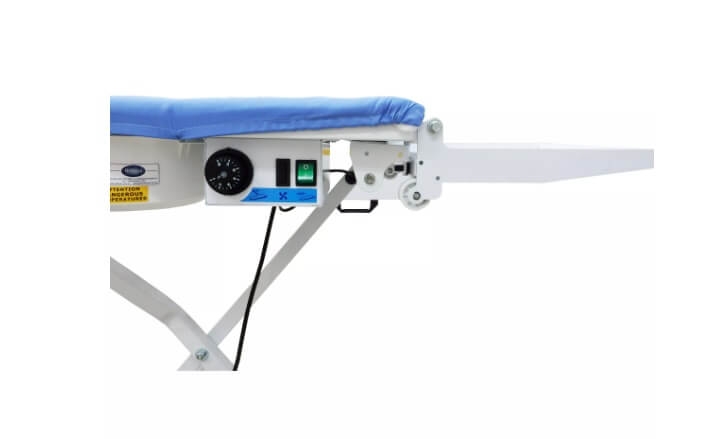 Stół prasowalniczy Comel Comelflex z niebieską powierzchnią roboczą, składany. Z boku widoczny panel sterujący z zegarem, przyciskiem włączania i kontrolką. Stelaż metalowy, stabilny. Wysuwana półka na boku.