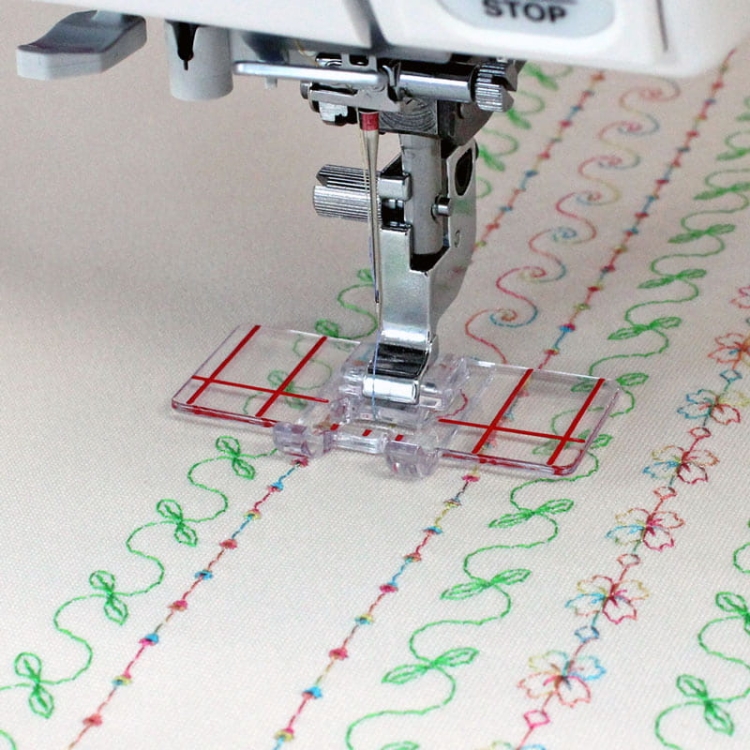 Stopka do równoległego prowadzenia ściegu Janome, przezroczysta, z czerwonymi liniami na powierzchni. Ujęcie z boku, pokazujące stopkę zamontowaną na maszynie do szycia, nad tkaniną z kolorowymi haftami w kształcie wstążek i kwiatów.