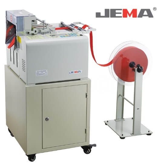 Urządzenie tnące JEMA JM-130L z automatycznym systemem, przeznaczone do cięcia średnich i grubych materiałów na zimno. Na zdjęciu widoczny jest sprzęt w kolorze szarym z czerwonymi elementami oraz stojak z rolką materiału.