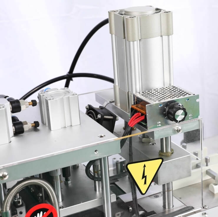Urządzenie do cięcia taśmy JEMA JM-150LR z automatycznym podajnikiem. Widoczny silnik, elementy metalowe w kolorze srebrnym, oznaczenie z symbolem ostrzegawczym o napięciu. Kąt ujęcia z boku, detal konstrukcji maszyny.