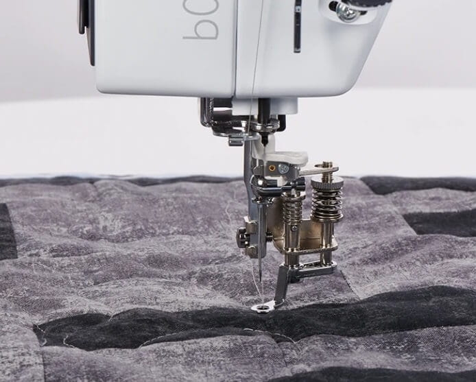 Maszyna do szycia BERNETTE 08 Straight Stitch w zbliżeniu, pokazująca stopkę i igłę, szyjącą przez warstwy materiału o szarym i czarnym wzorze. Elementy mechaniczne widoczne obok igły, co podkreśla precyzyjność urządzenia.