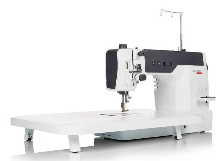 Maszyna do szycia BERNETTE 08 Straight Stitch w kolorze białym, z prostokątnym blatem. Widoczny mechanizm szyjący z igłą oraz panel sterujący z przyciskami. Ujęcie z boku, ukazujące detal konstrukcji i funkcjonalności urządzenia.