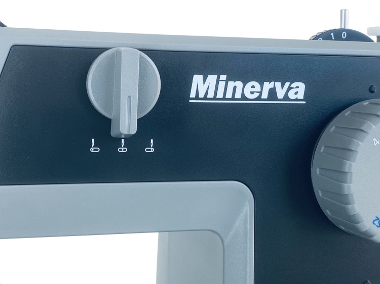 Panel sterujący maszyny do szycia Minerva HS1100 Heavy Duty, z logo Minerva. Widoczne pokrętło do regulacji oraz trzy kontrolki. Kolor szary z ciemnym tłem, z wyraźnymi oznaczeniami.
