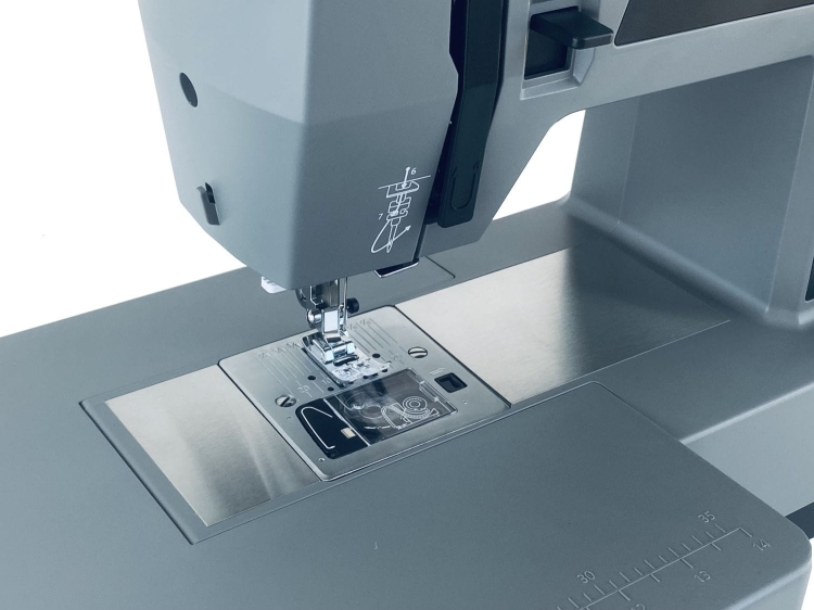 Maszyna do szycia Minerva HS1100 Heavy Duty z widocznym metalowym stółkiem, stopką i igłą. Ujęcie z góry, ukazujące panel sterujący oraz oznaczenia na obudowie. Główna część w kolorze szarym, z detalami w odcieniach metalicznych.
