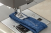 Maszyna do szycia Redstar RQ60 Heavy Duty z bliska, z igłą przeszywającą niebieski materiał denim. Widać metalową stopkę oraz panel z regulacją długości ściegu. Tło jest proste, co podkreśla detale maszyny.