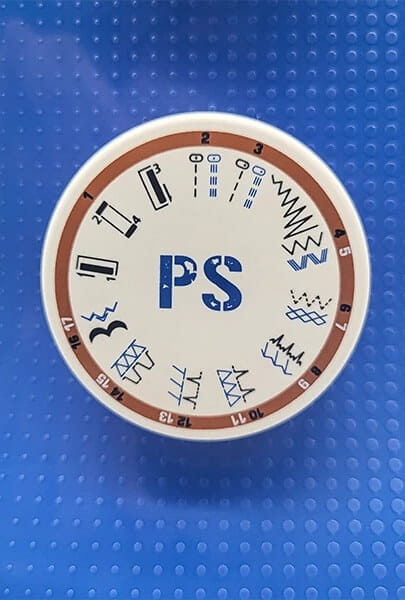 Zbliżenie na pokrętło maszyny do szycia Veritas Power Stitch Jeans. Białe tło z brązowym obrzeżem, widoczne symbole ściegów oraz oznaczenie PS w niebieskim kolorze. Pokrętło znajduje się na niebieskim tle.