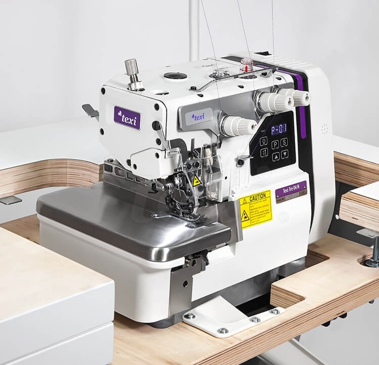 Overlock TEXI TRE 04 N PREMIUM 3-nitkowy, z widocznym metalowym blatem, panel sterujący z wyświetlaczem, białą obudową i logo texi. Maszyna stoi na stoliku, otoczona drewnianymi elementami. Wyposażona w cztery uchwyty do nici i ostrze.