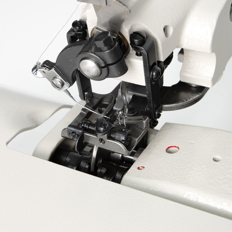 Podszywarka TEXI ACCURA 2.0 PREMIUM w zbliżeniu, ukazująca mechanizm szyjący, igłę i chwytacz. Elementy metalowe w kolorze czarnym z wyraźnymi detalami. Widoczna także część materiału oraz otwór w podłożu. Całość na jasnym tle.