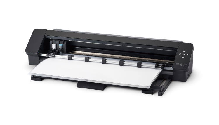 Ploter tnący Silhouette Cameo Pro MK II w kolorze czarnym, z widocznym białym arkuszem papieru na stole. Urządzenie ma przyciski sterujące po prawej stronie oraz dwa wkłady w kolorach niebieskim i czarnym po lewej stronie.