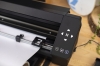 Ploter tnący Silhouette Cameo Pro MK II w kolorze czarnym, widoczny z boku. Na górze znajduje się panel sterujący z przyciskami nawigacyjnymi i ikoną Bluetooth. Obok widać rolkę z białym papierem, a tło jest w naturalnym otoczeniu z drewnianym blatem.