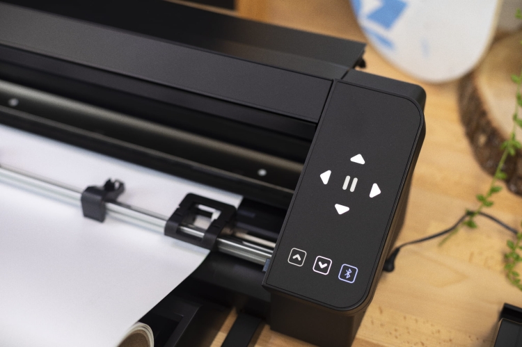 Ploter tnący Silhouette Cameo Pro MK II w kolorze czarnym, widoczny z boku. Na górze znajduje się panel sterujący z przyciskami nawigacyjnymi i ikoną Bluetooth. Obok widać rolkę z białym papierem, a tło jest w naturalnym otoczeniu z drewnianym blatem.