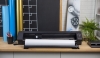 Ploter tnący Silhouette Cameo Pro MK II w kolorze czarnym, umieszczony na drewnianym blacie. W tle widoczne kolorowe rolki folii i zielona roślina. Ustawiony w pozycji otwartej, z widoczną rolką papieru. Panel sterujący z przyciskami po prawej stronie.