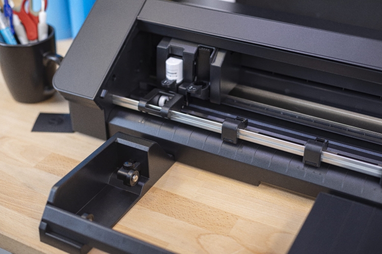 Ploter tnący Silhouette Cameo Pro MK II w kolorze czarnym, widok z boku, ukazujący otwartą komorę na narzędzia oraz elementy robocze. Produkt stoi na drewnianym stole, w tle widać kubek z przyborami biurowymi.