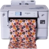 Drukarka Brother HL-JF1 PrintModa Studio w kolorze białym, z wyświetlaczem dotykowym, z rolką papieru w różowe kwiaty na jasnym tle, wydobywająca wzór. Produkt stoi na płaskiej powierzchni, z widocznym logo na obudowie.