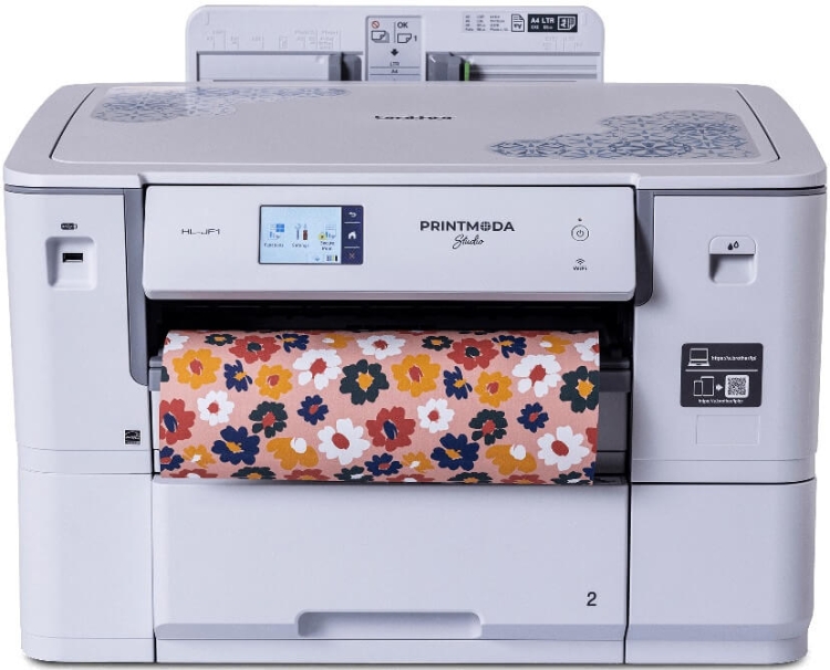 Drukarka Brother HL-JF1 PrintModa Studio w kolorze białym z kolorowym papierem w kwiatowy wzór, z wyświetlaczem dotykowym i przyciskami. Ujęcie frontalne, urządzenie stoi na podłożu, z widoczną rolką papieru.