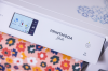 Drukarka Brother HL-JF1 PrintModa Studio w kolorze białym z wyświetlaczem dotykowym, widoczna funkcja Wi-Fi. Na tle kolorowego wzoru tkaniny, z logo PrintModa Studio. Produkt stoi na płaskiej powierzchni, z widocznymi przyciskami i ikonami na panelu.