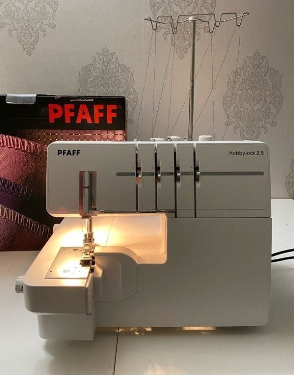 Owerlok PFAFF HOBBYLOCK 2.5, 4-nitkowy, w kolorze białym, z podświetleniem roboczym. Widoczny w frontalnej pozycji z wbudowanym uchwytem na nici i logo PFAFF. Tło w stonowanej kolorystyce z wzorem.