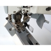 Zbliżenie na mechanizm autolapu ramieniowego SIRUBA D007S-460-02ET/AW1. Widoczne metalowe elementy, w tym igła, stopka i prowadnik materiału, na błyszczącej, stalowej podstawie. Przycisk i sprężyna w tle. Tło jasne, podkreślające detale maszyny.