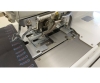 Dziurkarka odzieżowa SIRUBA BH-9820-01 z automatycznym mechanizmem, widok z góry na stół roboczy, metalowe elementy, dwie nitki, szare tło materiału z kolorowym haftem. Widać igłę oraz mechanizm naciągu.