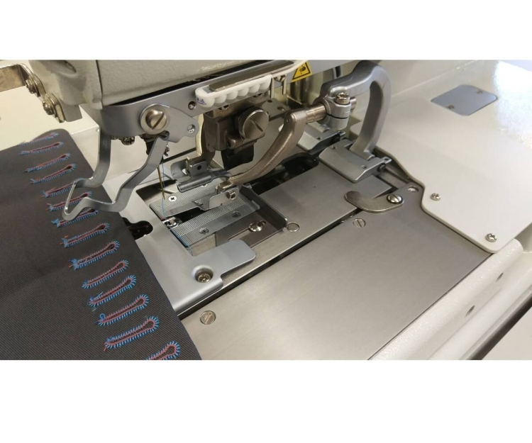 Dziurkarka odzieżowa SIRUBA BH-9820-01 z automatycznym mechanizmem, widok z góry na stół roboczy, metalowe elementy, dwie nitki, szare tło materiału z kolorowym haftem. Widać igłę oraz mechanizm naciągu.