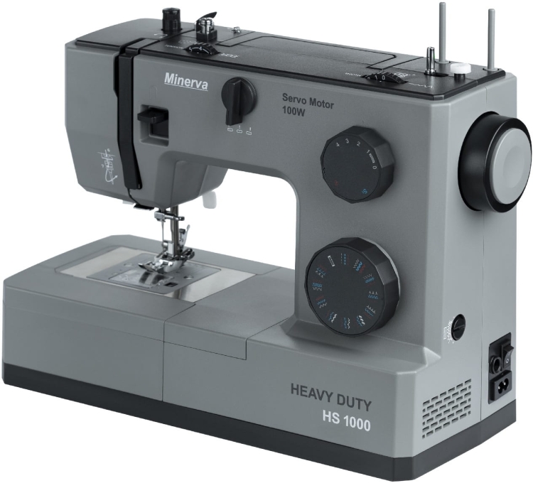 Maszyna do szycia Minerva HS1000, mechaniczna, w szarym kolorze, z widocznymi pokrętłami regulacji i oznaczeniem HEAVY DUTY. Ujęcie frontalne, pokazujące stół roboczy i igłę, zasilanie z prawej strony.