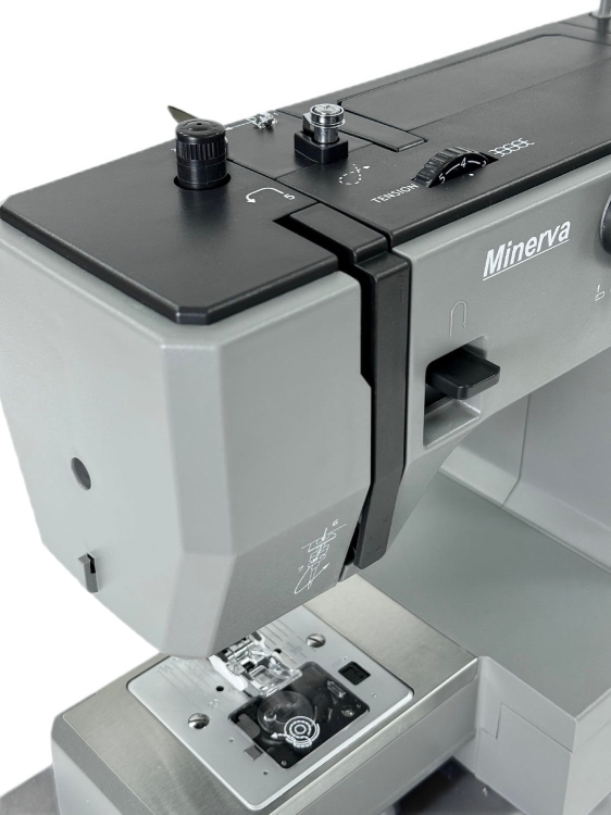Maszyna do szycia Minerva HS1000 w kolorze szarym, z widocznymi pokrętłami do regulacji napięcia i długości ściegu oraz metalową płytką z igłą i stopką. Ujęcie z góry, pokazujące detale mechaniczne i przyciski.