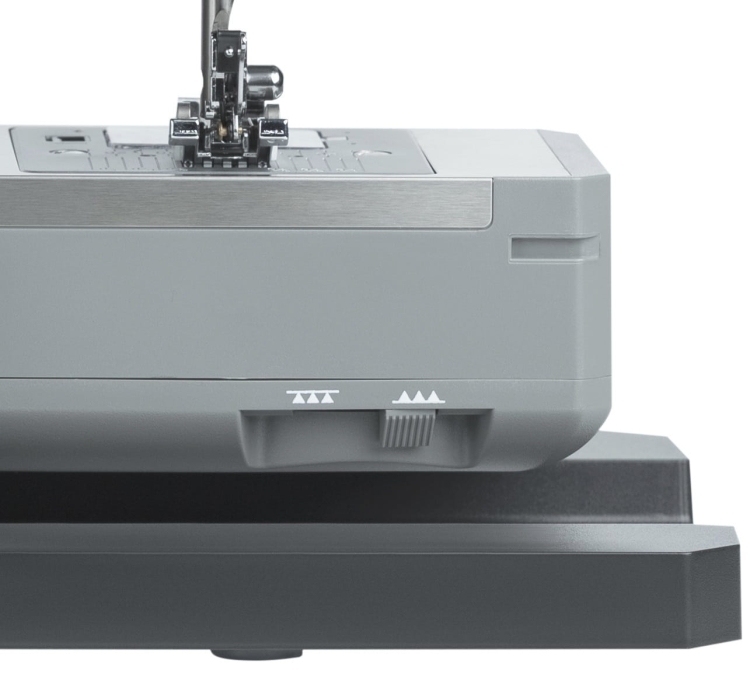 Maszyna do szycia Minerva HS1000 w kolorze szarym, widok z boku. Na przednim panelu znajdują się oznaczenia do regulacji ściegów oraz przestrzeń na stopkę. Produkt ustawiony na czarnej podstawie, z metalowym mechanizmem szyjącym widocznym na górze.