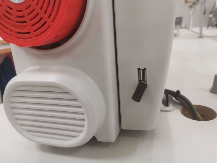 Zbliżenie na bok stebnówki SIRUBA DL7200C-BH1-16Q, z widocznym czerwonym wentylatorem i portem USB. Maszyna ma białą obudowę, z prostokątną kratką wentylacyjną u dołu. Widoczne są także kable zasilające.