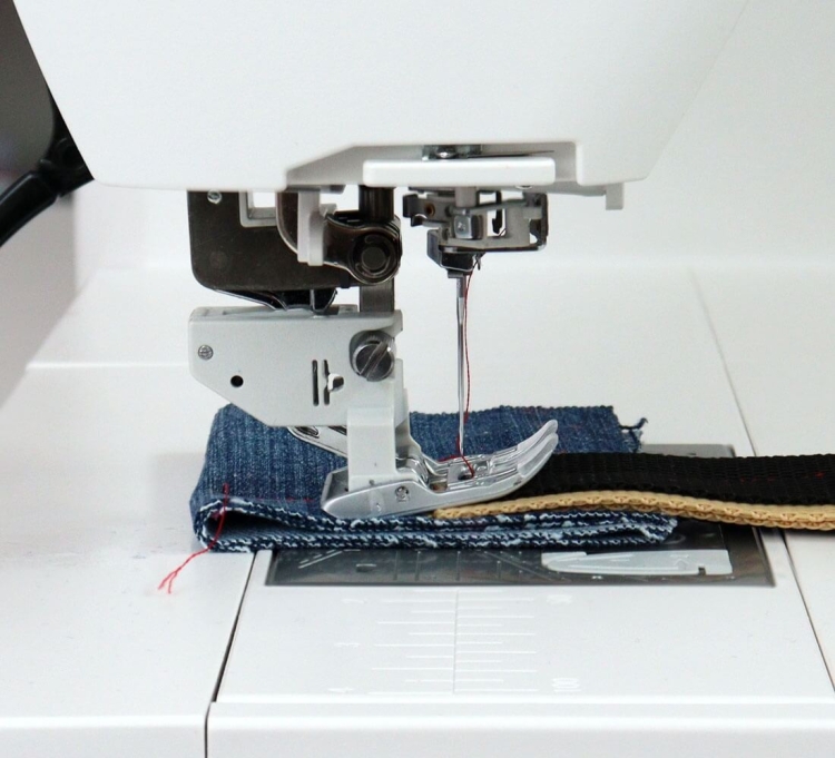 Maszyna do szycia Janome Skyline S6 z widoczną stopką do szycia na warstwie materiału denimu i taśmie, z igłą w pozycji gotowości. Ujęcie z boku, pokazujące detale mechanizmu i materiału na stole roboczym.