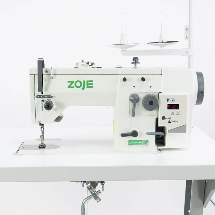 Zygzak ZOJE ZJ20U93-BD to przemysłowa maszyna do szycia w kolorze białym, z widocznym logo ZOJE. Ujęcie frontalne, pokazujące stół roboczy oraz mechanizm szycia z pokrętłami regulacyjnymi. W tle dwa stojaki na nici.