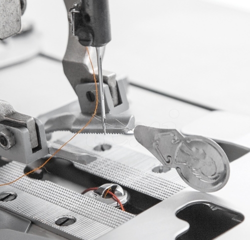 Nawlekacz nici do igły w zestawie 2 sztuki, metalowy, ukazany z bliska przy maszynie do szycia. Widać igłę z nawleczoną nicią w kolorze pomarańczowym oraz mechanizm nawlekania. Tło to elementy maszyny oraz metalowa stopka.