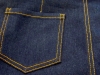 Zbliżenie na tylną kieszeń dżinsów, wykonane z ciemnoniebieskiego materiału, z widocznymi żółtymi szwami. Kształt kieszeni jest prostokątny, z delikatnymi przeszyciami na brzegach. Tło jednolite, co podkreśla detal kieszeni.