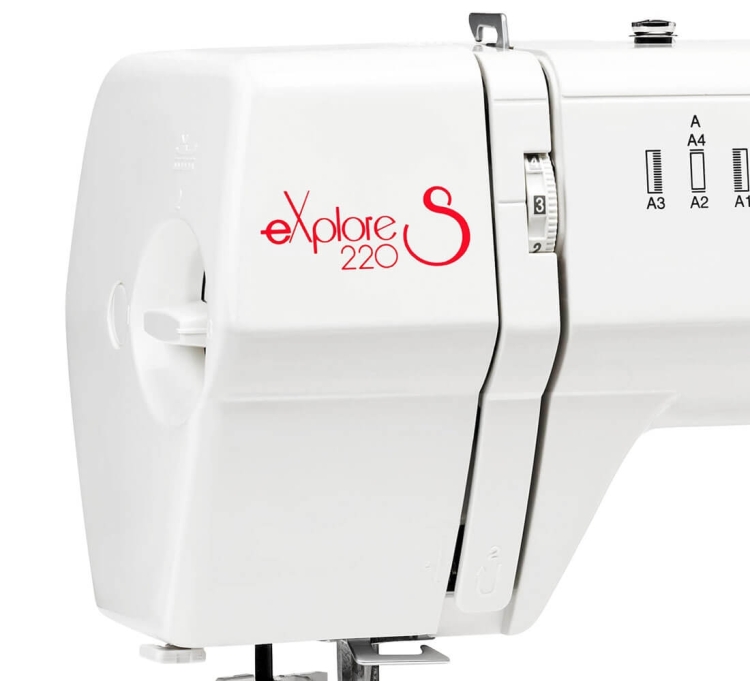 Maszyna do szycia Elna eXplore 220S w kolorze białym. Widoczne logo eXplore 220 w czerwonym kolorze, pokrętło regulacji oraz znaki wskazujące różne ustawienia. Produkt ustawiony w pozycji frontalnej, z widocznym mechanizmem szycia.