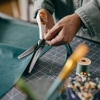 Nożyczki krawieckie Fiskars 1070024 ZygZak SoftGrip o długości 23 cm, z ząbkowanym ostrzem, używane do cięcia zielonego materiału. Ujęcie z boku, ręka trzymająca nożyczki, na tle maty do szycia z widocznymi akcesoriami.