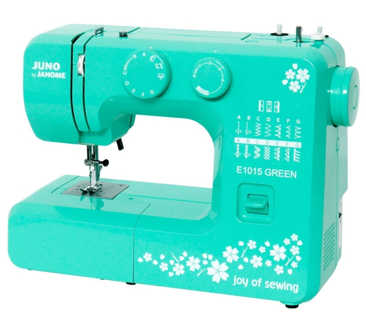 Maszyna do szycia Janome JUNO E1015 w kolorze zielonym. Widoczna jest szeregowa skala ściegów oraz pokrętła do regulacji. Na obudowie biała dekoracja kwiatowa. Ujęcie frontalne, maszyna stoi na płaskiej powierzchni.