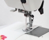 Maszyna do szycia Janome HD 523 EASY JEANS w zbliżeniu, pokazująca stopkę do szycia na białym materiale z kolorowym wzorem. Widoczne są nici oraz mechanizm szyjący z uchwytem. Tło proste, białe.