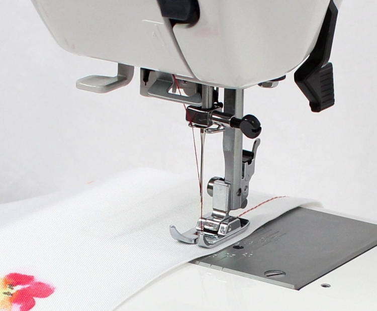 Maszyna do szycia Janome HD 523 EASY JEANS w zbliżeniu, pokazująca stopkę do szycia na białym materiale z kolorowym wzorem. Widoczne są nici oraz mechanizm szyjący z uchwytem. Tło proste, białe.