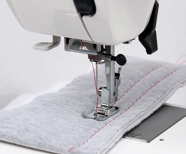 Maszyna do szycia Janome HD 523 EASY JEANS, widok z bliska na stopkę oraz igłę szyjącą, z szarym materiałem z widocznym czerwonym ściegiem. Elementy maszyny są w kolorze białym i metalicznym, z ergonomicznym uchwytem oraz pedałem.