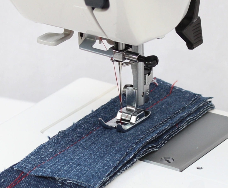 Maszyna do szycia Janome HD 523 EASY JEANS z widocznym stopkiem i igłą przyszywającą kilka warstw niebieskiego denimu. W tle elementy maszyny, w tym pokrętła i panel sterujący. Stół maszyny w jasnym kolorze, z detalami szycia w czerwonym włóknie.