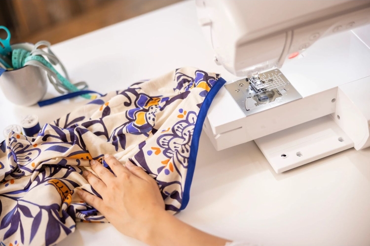 Maszyna do szycia Brother Inventio NV1045SE, widoczna z boku, z materiałem w kwiatowy wzór w odcieniach beżu, niebieskiego i pomarańczowego, pod ręką użytkownika. Przypinacz i akcesoria do szycia w tle na białym stole.