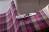 Maszyna do szycia Brother Inventio NV1350 w zbliżeniu na stopkę do szycia, używana na tkaninie w kratkę w odcieniach różu, beżu i fioletu. Widoczne detale: biały mechanizm stopki, różowa podkładka. Tło jasne, neutralne.