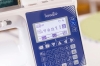 Maszyna do szycia Brother Inventio NV1850Q z widocznym wyświetlaczem LCD, który pokazuje logo Inventio. Panel sterujący z przyciskami i symbolami szycia. W tle widoczna szpula z niebieską nicią. Produkt na białym tle.