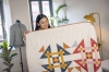 Kobieta trzyma patchworkowy koc w kolorach czerwonym, niebieskim, pomarańczowym i żółtym, z geometrycznym wzorem. W tle widoczna maszyna do szycia oraz stojący manekin. Oświetlenie naturalne, z oknami w tle, rośliny w aranżacji wnętrza.