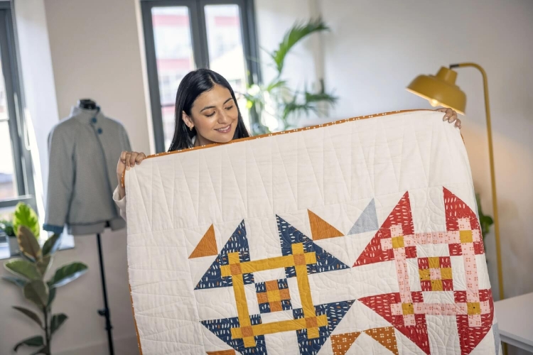 Kobieta trzyma patchworkowy koc w kolorach czerwonym, niebieskim, pomarańczowym i żółtym, z geometrycznym wzorem. W tle widoczna maszyna do szycia oraz stojący manekin. Oświetlenie naturalne, z oknami w tle, rośliny w aranżacji wnętrza.